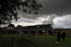 'Flying Scotsman' leaves Wansford under a stormy sky, watched by two ladies.